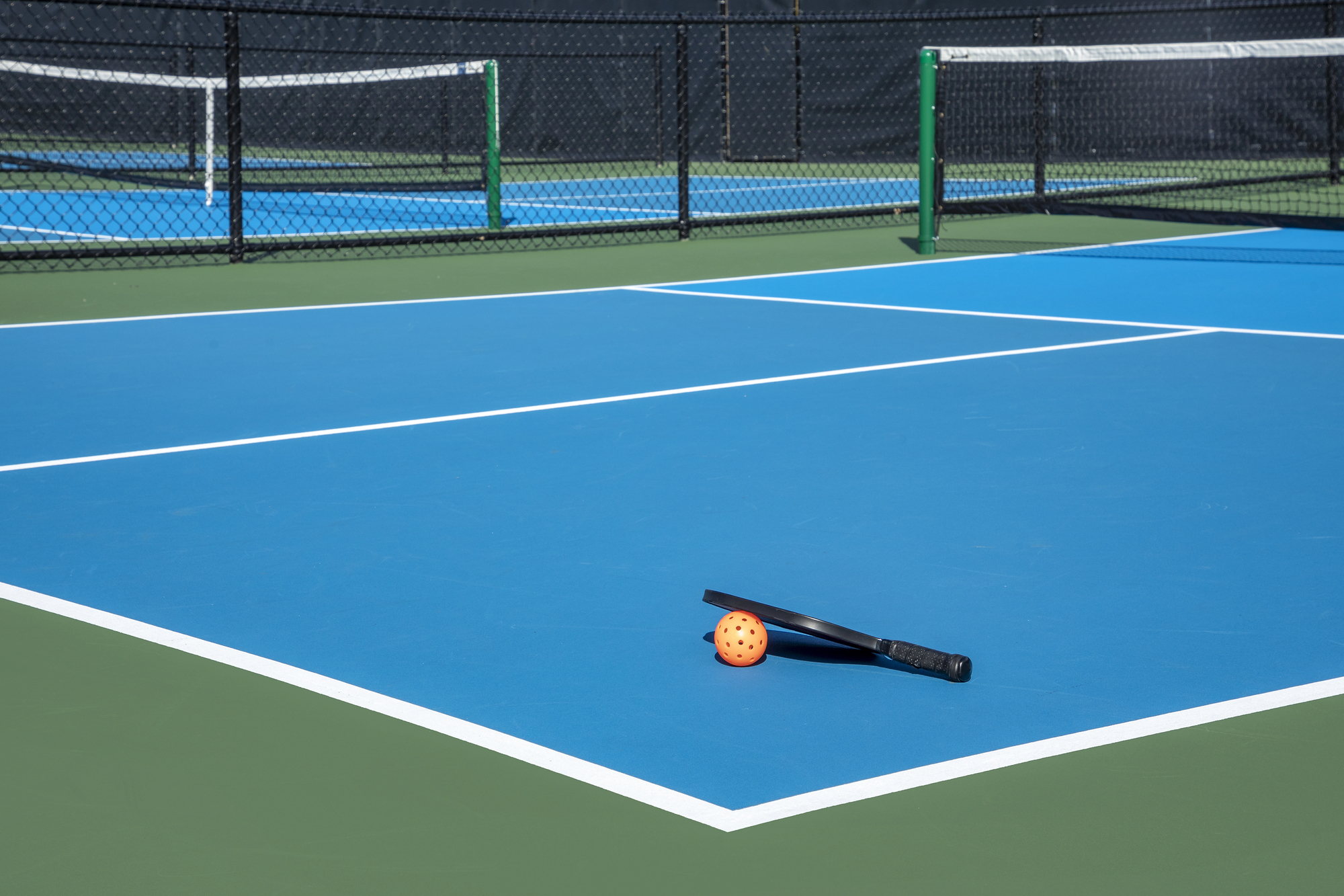 View of a new pickleball complex with a paddle and orange ball on blue and green courts in a suburban setting during early spring.