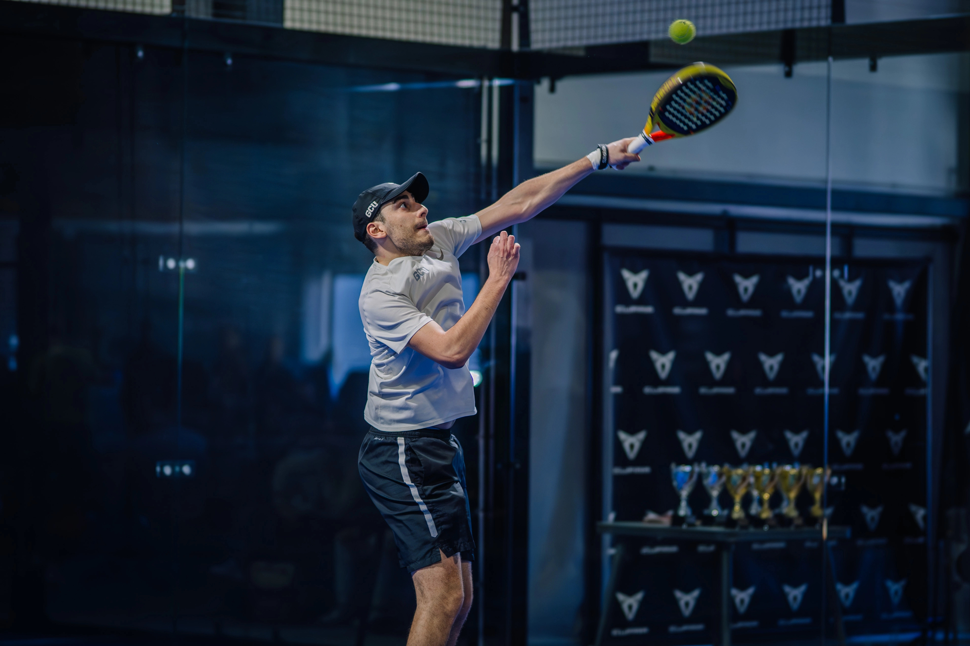 Riga, Latvia - March 3, 2024 - A man in a cap and sportswear reaches for an overhead shot in a padel game, with trophies and an audience visible in the background — Photo by sarelainens@gmail.com